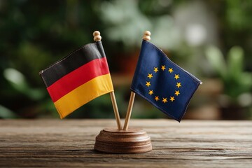 Two flags Germany the European Union stand sidebyside on a wood base atop a wooden table against a blurred green leafy background
