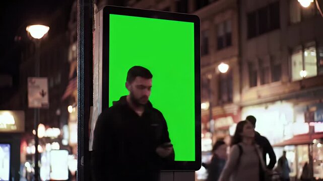 Man Engaged with Smartphone in Front of a Large Green Screen Digital Billboard on a Busy City Street at Night, Ideal for Advertising Mockups and Ur...