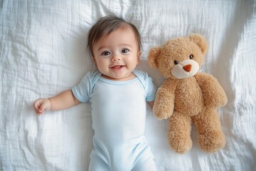 Happy baby lying on white bed next to a soft brown teddy bear, smiling joyfully with bright eyes and wearing a light blue onesie