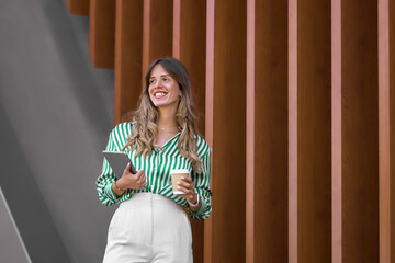 Young pretty smiling businesswoman in green striped shirt with tablet and coffee to go happily looking away on street