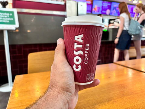 Alicante, Spain - August 26, 2025: Hand holding a red Costa Coffee cup in a cafe, with blurred customers in the background, showcasing a vibrant coffee shop atmosphere
