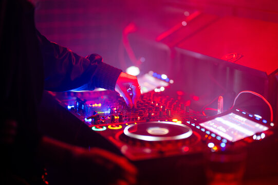 DJ adjusting mixer controls on an illuminated club deck bathed in red and blue stage lights.