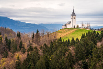 Fototapeta premium Hermosa postal de la Iglesia de Jamnik, en Eslovenia, ubicada en la cima de una colina rodeada de bosques, un día de otoño