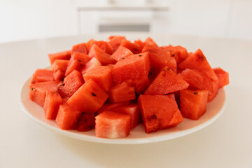 A plate filled with vibrant watermelon cubes sits on a white countertop in a sunny kitchen. The pieces show juicy red flesh and are ready to be enjoyed as a refreshing snack
