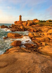 Scenic view of the Ploumanac'h lighthouse on the Pink Granite Coast in Brittany, France. The rugged coastline with unique red granite rock formations is illuminated by warm sunset light. 