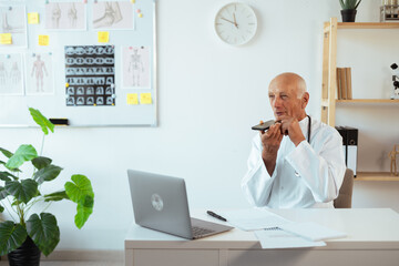 Senior elderly male doctor recording audio message to a patient using smartphone mic working in clinic or hospital.
