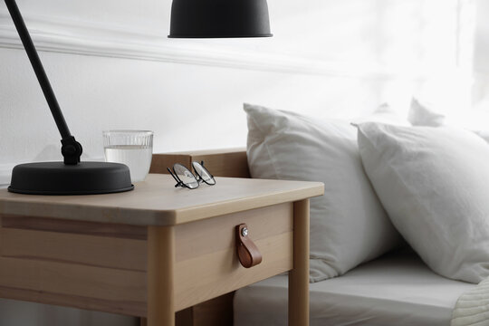 Bedside table with lamp, notebook, glasses and glass of water near bed indoors, closeup - Powered by Adobe