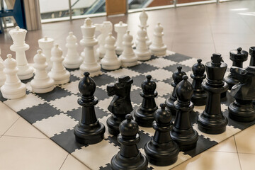 A large chess board is displayed indoors, featuring oversized black and white pieces. Players prepare to engage in a thoughtful game. Natural light illuminates the setting
