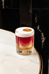 Colorful layered cocktail with foam served in a crystal glass, garnished with a dried citrus slice. Elegant drink presentation on a marble table in a stylish modern bar