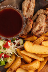 op view of a delicious meal with grilled meat, golden potato wedges, fresh vegetable salad, and a glass bowl of red sauce on a wooden plate, served on a stone background