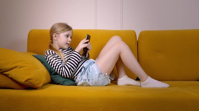Relaxed preteen girl enjoying mobile gaming while lying comfortably on vibrant yellow sofa, immersed in digital entertainment at home