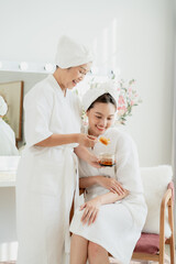 Senior asian woman feeding honey with spoon to teenage daughter in white spa robes sitting together, expressing warm love bonding and healthy skincare moment in home wellness lifestyle concept