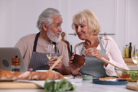 Elderly couple with laptop cooking together and drinking white wine at table indoors