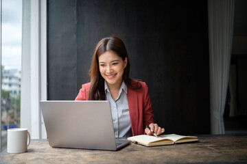 Businesswoman in a red blazer writing in notebook while working on a laptop at a wooden desk by the window