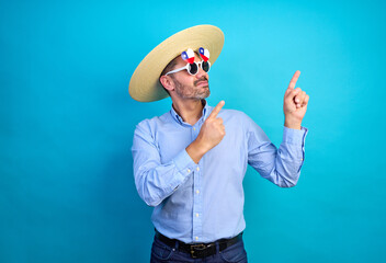 Studio portrait of man wearing straw hat chupalla, funny Chile flag glasses, and light blue shirt pointing up with index fingers against a blue background, national holiday and patriotism celebration.
