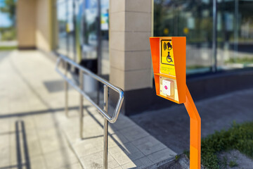Side view of accessibility button with sign stands on city sidewalk. Sign wheelchair symbol, bell icon, and Braille text, aiding disabled individuals. Glass building with stairs on background.