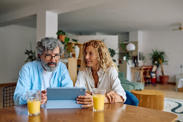Happy mature couple is using digital tablet and drinking orange juice while sitting at dining table at home