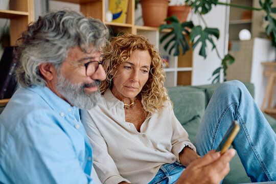 Senior couple sitting together on a cozy sofa at home, looking at a smartphone and enjoying their time connecting online - Powered by Adobe