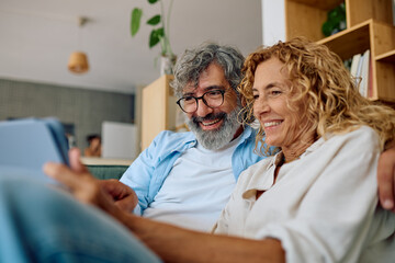 Obraz premium Cheerful retired couple enjoying modern technology, browsing internet or watching videos on digital tablet, spending leisure time together in cozy living room