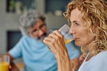 Mature woman savoring a refreshing glass of orange juice at home, with her husband enjoying the morning in the background