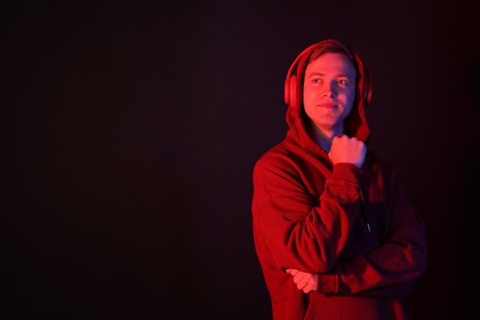 Handsome man wearing hoodie and headphones in neon lights against black background. Space for text