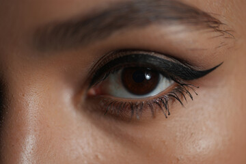 Obraz premium Close-up of a woman's dark brown eye with dramatic winged eyeliner, showcasing rich skin tones and intense gaze.