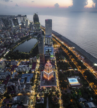 Aerial view of illuminated buildings casting a warm glow against the darkening skyline, contrasting with the cool blues of the sea, Batumi, Adjara, Georgia.