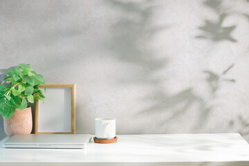 Minimalist workspace with a closed laptop, coffee cup, potted plant, and wooden photo frame on a white desk against a light wall.