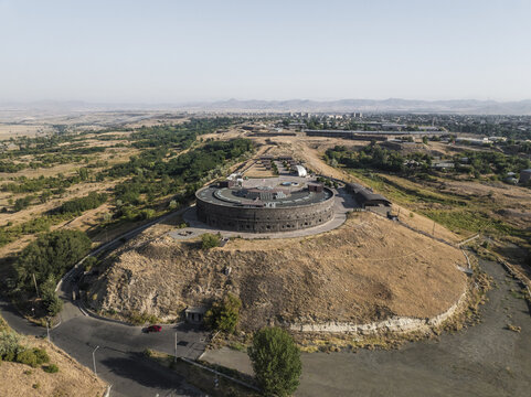 Aerial view of the stark, circular Sev Berd fortress, a dark monument atop a hill against the vast, sun-baked landscape, Gyumri, Shirak Province, Armenia.