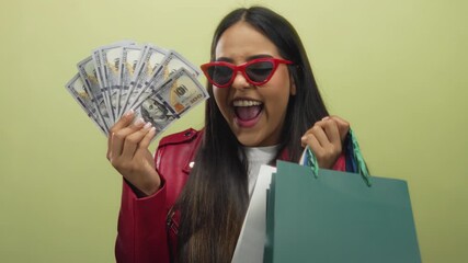 Woman holding shopping bags and us dollars joyfully smiles in red jacket and sunglasses against an isolated yellow background, capturing the essence of shopping and wealth.