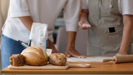 Freshly baked artisanal bread on a wooden board with a couple preparing dough in the background.