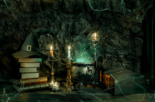 Halloween background. Dark witch cauldron with a magic witch's potion, books, chest, conical hat. Witchcraft paraphernalia and accessories among candles on a table in a room or cave with stone walls.