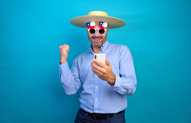 Enthusiastic chilean man expressing excitement with smartphone, wearing straw hat chupalla, chilean flag glasses, and clenched fist, celebrating national pride on vibrant blue backdrop