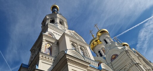 A grand Orthodox church with white walls and gleaming golden onion domes stands against a bright blue sky streaked with thin white clouds. Mosaics of saints adorn the facade.