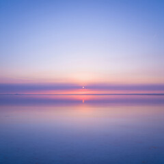 Fototapeta premium Serene water reflecting the pastel sky with a faint sun on the horizon at the end of the day
