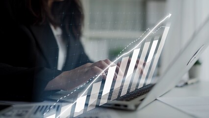 A businesswoman engages in typing on her laptop while an ascending bar graph appears digitally overlaid, symbolizing growth and success in a modern office setting. Copula