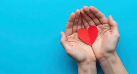 A pair of hands holding a red paper heart against a bright blue background studio shot