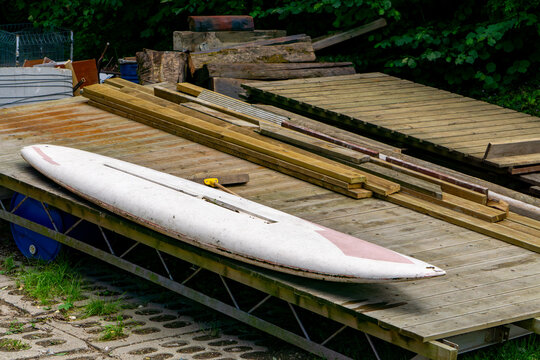 Old surfboard lying on a wooden pier next to a stack of planks and building materials.