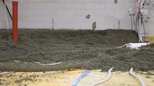 Liquid foam concrete screed flows from a hose, covering underfloor heating pipes on yellow sheeting in an indoor construction site with white brick walls.