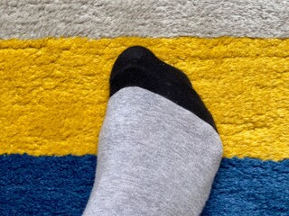 Close view of foot wearing black and grey sock resting on colorful rug, with yellow, blue and grey horizontal stripes on the textured carpet