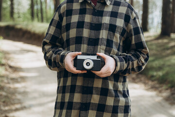 A man in a checkered shirt holds a film camera in his hands in the forest. Old film camera in a male hand on a blurred background of nature. The concept of travel, tourism and analog photography.
