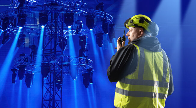 Stage technician in helmet and safety vest using radio while inspecting concert lighting rig with blue spotlights. Entertainment, Event Production, Stage Lighting.