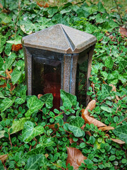 Memorial candle among wet green grass and autumn fallen leaves. Brass construction covered with patina, with thick glass. Installed in cemetery.