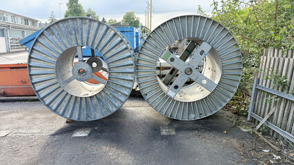Two large industrial metal cable reels on asphalt, front view outdoors: corrugated steel spools standing upright near fence and containers