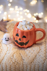Halloween still life with pumpkin cup filled with chocolate and marshmallows, little ghost and string lights on a white wool sweater. Festive, cozy  background with copy space. Vertical shot.