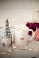 Christmas still life with white heart cup filled with chocolate and marshmallows, gnome doll and string lights. Festive, cozy  background with copy space. Vertical shot.