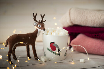 Christmas still life with robin cup filled with chocolate and marshmallows, deer doll and string lights. Festive, cozy  background with copy space.