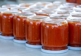 Rows of freshly canned homemade tomato sauce in glass jars, cooling upside down on a table. Traditional food preservation process for making winter preparations.