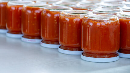 Rows of freshly canned homemade tomato sauce in glass jars, cooling upside down on a table. Traditional food preservation process for making winter preparations.