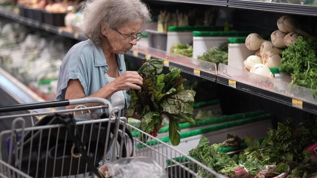 An elderly woman browses through fresh vegetables at a grocery store, carefully selecting leafy greens and placing them in her shopping cart during the day.